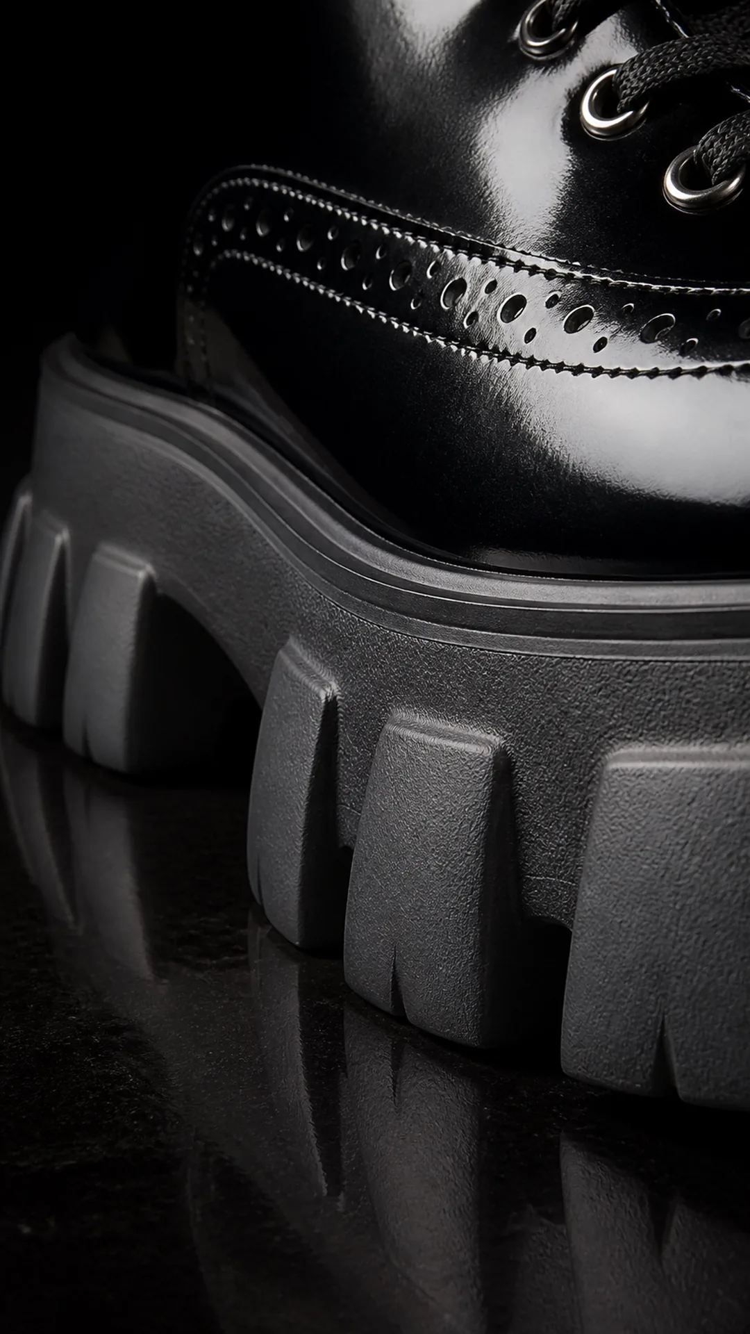 Extreme macro of the lugged platform sole of a black patent leather combat boot, raking light picking out each tread block against a dark floor.